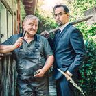 Ein Handwerker lächelt mit Zange, neben ihm steht ein ernster Mann im Anzug mit Spaten, vor einer Holzwand.