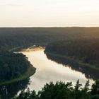 Sonnenuntergang über einem Fluss inmitten eines dichten Waldes, mit ruhigem Wasser und weitläufigen Bäumen.