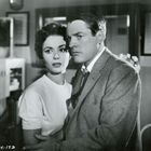 Text "5504-19D". Eine Frau in einem weißen Oberteil und ein Mann in einem Anzug stehen nah beieinander im Raum.