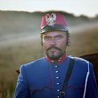 Ein Mann in einer blauen Uniform mit rotem Hut und Abzeichen steht auf einem Feld, im Hintergrund ein unscharfer Hügel.
