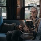 Eine Frau sitzt auf einem Sofa und liest ein Buch, umgeben von Kissen. Große Fenster im Hintergrund zeigen Bäume draußen.