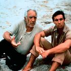 Zwei Männer sitzen im Sand. Der Ältere gestikuliert, während der Jüngere nachdenklich schaut. Beide sind lässig gekleidet.