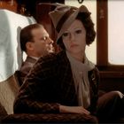 Eine elegant gekleidete Frau in einem Zugabteil, mit Hut und Schal, sitzt gedankenversunken, im Hintergrund ein Mann.