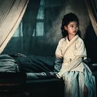 Ein Kind in traditioneller Kleidung sitzt besorgt neben einer Person auf einem Bett, im spärlich beleuchteten Raum.