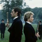 Zwei Männer stehen Rücken an Rücken auf einer Wiese, jeweils eine Pistole in der Hand haltend, als würden sie ein Duell austragen.