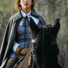 Eine Frau in historischer Kleidung reitet auf einem schwarzen Pferd, schaut konzentriert nach vorne.