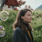 Eine Frau mit braunem Haar steht im Profil in einer grünen Wiese, umgeben von schwebenden Seifenblasen.