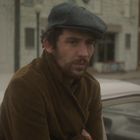Ein Mann mit braunem Mantel lehnt an einem Auto, trägt eine graue Schiebermütze, städtische Umgebung im Hintergrund.