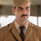 Ein Mann trägt eine beige Uniform mit Krawatte und Mütze, schaut ernst, mit einem Schnurrbart.
