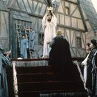 Eine Person in einem weißen Gewand hält eine Krone hoch; umgeben von applaudierenden Menschen in mittelalterlicher Kleidung.