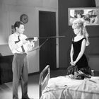 Eine Uhr zeigt 10:10. Ein Mann zielt mit einem Gewehr auf eine Frau. Beide befinden sich in einem Raum mit einem Bett.