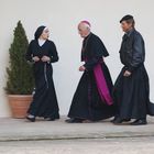 Ein Bischof, eine Nonne und ein Priester in schwarzem Gewand gehen nebeneinander an einem Zypressenbaum vorbei.