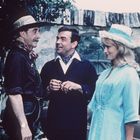 Drei Personen stehen nebeneinander: ein Mann mit Hut, ein lachender Mann und eine Frau in blauem Kleid und Hut vor einem Auto.