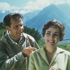 Ich kann keine Texte im Bild erkennen. Ein Mann und eine Frau stehen vor einer malerischen Berglandschaft.