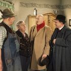 Vier ältere Männer in traditioneller Kleidung stehen in einem rustikal eingerichteten Raum zusammen.