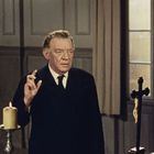 Ein alter Mann in Anzug hebt die Hand, neben ihm eine brennende Kerze und ein Kreuz vor hellen Fenstern.