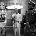 Drei Männer stehen draußen: zwei in glänzenden Uniformen, einer mit einem weißen Regenschirm in der Mitte.