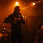 Ein Sänger performt vor einer Band in einem orange beleuchteten Raum. Ein Schlagzeuger spielt im Hintergrund.