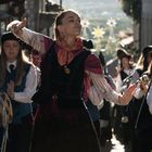 Eine Gruppe von Menschen, vor allem Frauen, in traditioneller Kleidung, geht eine sonnenbeschienene Straße entlang. Einige tragen Tamburine.