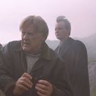 Zwei ältere Männer, einer hält ein Fernglas in felsiger, nebliger Landschaft. Der vordere Mann schaut angespannt.