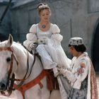 Eine Frau in weißer Kleidung sitzt auf einem weißen Pferd, ein Mann in aufwendigem Kostüm hilft ihr elegant.