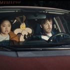 Eine Frau, ein Mann und eine Cartoonfigur sitzen in einem Auto. Die Frau trägt eine braune Jacke.