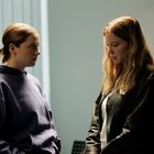 Zwei Frauen stehen sich gegenüber, die linke trägt einen blauen Hoodie, die rechte hat langes blondes Haar und trägt Schwarz.