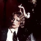 Eine Frau mit blonden Locken, elegant gekleidet, hält eine Zigarette und schaut nachdenklich zur Seite.