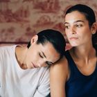 Zwei Menschen, eine Person lehnt den Kopf auf die Schulter der anderen. Hintergrund mit Rot-weißem Muster.