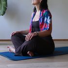 Eine Frau sitzt im Schneidersitz auf einer Yogamatte. Sie trägt einen bunten Schal mit Blumenmuster.