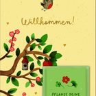 „Willkommen!“, „Pflanze deine Wünsche“. Ein Vogel fliegt über einen blühenden Baum mit Vogelhaus.