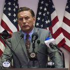 Ein Mann spricht vor Mikrofonen. Text auf Mikrofonen: „NEWS“. Hintergrund: Mehrere US-Flaggen.