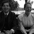 Zwei lächelnde Menschen in einem Wagen auf einem ländlichen Weg, umgeben von hohen Bäumen und Feldern, in Schwarz-Weiß.