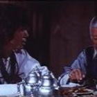 Zwei Personen in traditioneller Kleidung essen an einem Tisch; eine mit langem Haar, die andere mit Brille und grauem Haar.