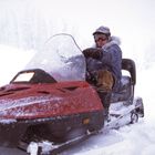 Ein Mensch fährt ein rotes Schneemobil im Schneesturm, ein weiteres Schneemobil im Hintergrund.