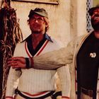 Zwei Männer, einer mit grünem Hut, lächeln nach oben. Beide tragen weiße Strickjacken mit rotem Streifen.