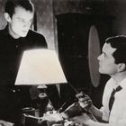 "JS-0-36" in der Ecke. Zwei Männer, einer steht, einer sitzt, sprechen an einem Tisch mit einer Lampe.