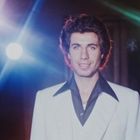 Kein Text vorhanden. Ein Mann in einem weißen Anzug mit schwarzem Hemd, vor bunten Lichtern in einer Disco-Atmosphäre.