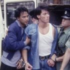 Ein Mann in Handschellen wird von zwei Polizisten in Uniform festgehalten; ein weiterer Mann steht daneben.