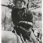 Ein Mann in Cowboy-Kleidung sitzt auf einem Pferd. Er trägt einen Hut und Handschuhe, umgeben von Bäumen im Hintergrund.