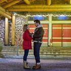 Ein Paar steht vor einem Gebäude mit roten Türen und Weihnachtlichter, umarmt sich und trägt Winterkleidung.