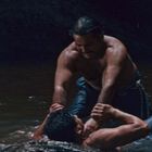 Zwei Männer kämpfen in einem flachen Fluss. Einer hält den Kopf des anderen fest. Im Hintergrund steht ein weiterer Mann im Wasser.