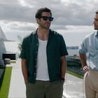 Zwei Männer stehen auf einer Terrasse mit Blick aufs Meer. Einer trägt ein dunkles Hemd, der andere ein helles.
