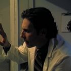 Ein Mann im weißen Kittel blickt durch eine Glaswand; daneben steht eine nachdenkliche Person.