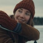 Eine Frau mit roter Strickmütze und Fäustlingen lächelt. Im Hintergrund winterliche Landschaft mit Wald und Schnee.
