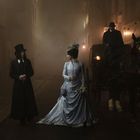 Unleserlicher, vernebelter Schriftzug im Hintergrund. Eine Dame in einem blauen Kleid und ein Herr in Schwarz auf nebliger Straße.