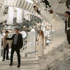 Mehrere weiße Treppen, darunter vier Personen, spiegelndes Umfeld. Atmosphäre surreal und verwirrend.