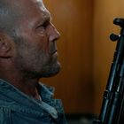 Ein Mann mit Bart in Jeansjacke hält ein Gewehr. Hintergrund ist holzgetäfelt, er wirkt nachdenklich und fokussiert.