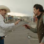 Zwei Männer auf einer leeren Straße gestikulieren miteinander. Links trägt einer einen Cowboyhut, rechts hat der andere eine Weste an.