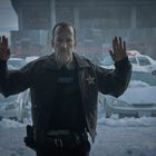 Ein Mann in Sheriff-Uniform steht mit erhobenen Händen vor einer Glastür, Schneelandschaft und Autos im Hintergrund.